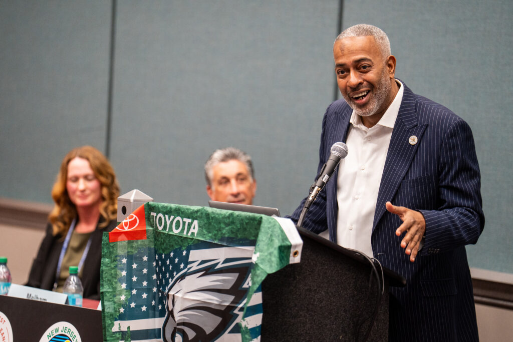 Speaker standing at podium speaking with other panelists in the background.
