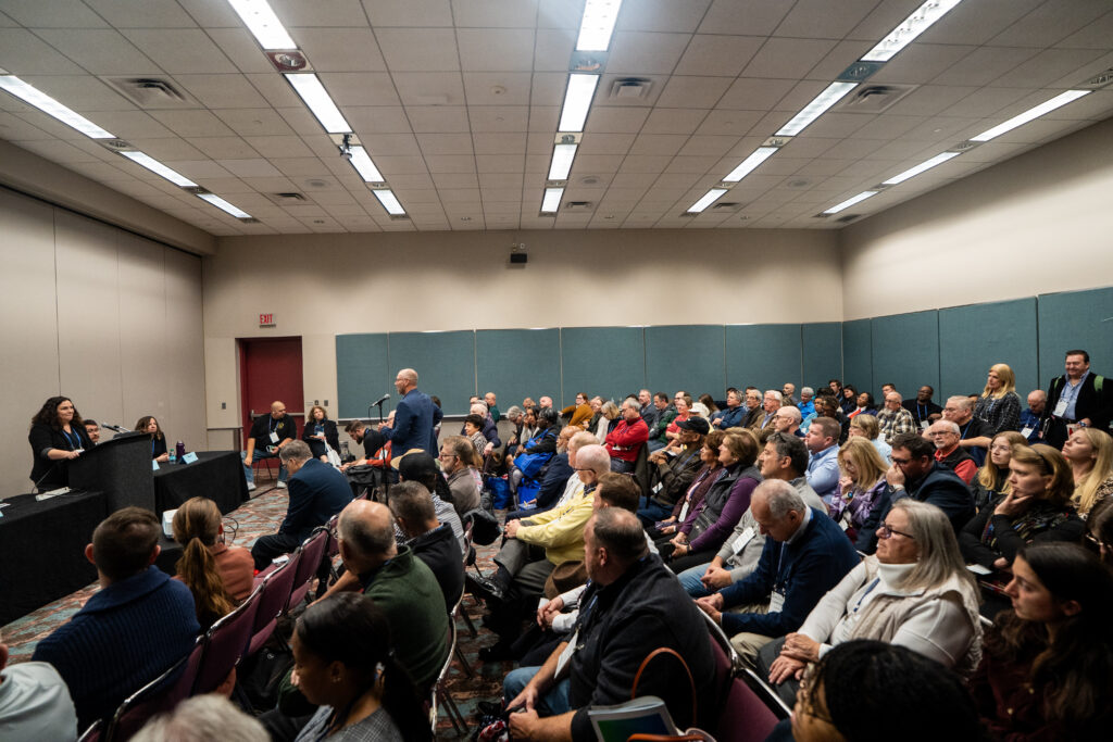 Full view of room with a large amount of people sitting and listening to a speaker panel.